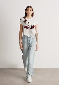 Sieviete iet iekštelpās, valkājot baltu T-kreklu, gaiši zilas džinsas, baltus sporta apavus un baltu džemperi ar sarkani melniem svītrām, kas sasiets pār pleciem.