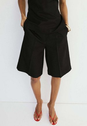 Persona que lleva pantalones cortos negros sueltos hasta la rodilla, camiseta negra, pulseras doradas y chanclas rojas, de pie contra una pared blanca lisa.