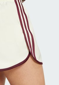 Bílé sportovní kraťasy s vínovými akcenty, zdobené třemi vertikálními pruhy po straně a zaobleným lemem. Hladký, lehký materiál.