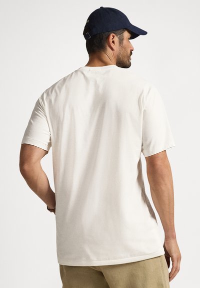 Μπλούζα από βαμβάκι σε κρεμ χρώμα με στρογγυλή λαιμόκοψη, κοντά μανίκια και άν ετη εφαρμογή, φορεμένη με ανοιχτόχρωμο χακί παντελόνι και καπέλο σε ναυτικό μπλε.