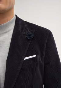 Blazer en velours côtelé bleu marine avec des rainures texturées, une poche poitrine simple affichant un mouchoir blanc et un accent de fleur en tissu sur le revers.