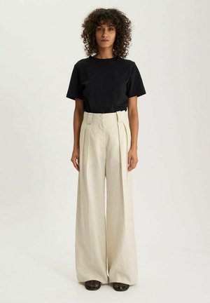 Femme aux cheveux bouclés debout portant un haut noir à manches courtes, un pantalon large crème taille haute et des chaussures tressées foncées sur un fond clair.