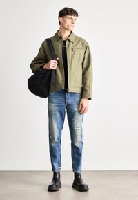 Olivengrønn jakke med en glidelåslomme, svart t-skjorte, slitt blå jeans, svart veske hengende over skulderen, og svarte skinn ankelsko.