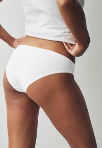 Vit bomull bikinibrief med en läkande textur, elastisk midja och vågig kant, bärs under en tigh-fitting vit linne.