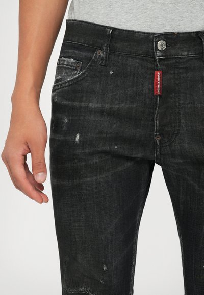 Pantalones vaqueros de mezclilla negra con un aspecto desgastado, que presentan detalles de desgarro. Incluyen una etiqueta de marca roja y bolsillos delanteros estándar.