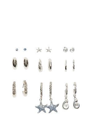 Dziewięć par srebrnych kolczyków na wystawie, w tym sztyfty, kółka, gwiazdy i wzory z fakturą, niektóre ozdobione niebieskimi kamieniami szlachetnymi.