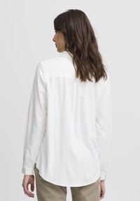 Camicia bianca a maniche lunghe con una texture liscia, polsini abbottonati e orlo curvo, che presenta un collo classico e un dettaglio di yoke singolo sul retro.