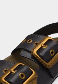 Μαύρο δερμάτινο σανδάλι με δύο ρυθμιζόμενους ιμάντες, που διαθέτουν μεγάλες υφασμένες χρυσές αγκράφες και αντίστοιχα χρυσά οφθαλμικά στοιχεία.