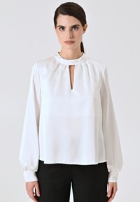 Weiße Bluse mit langen Ärmeln, rundem Ausschnitt und einem Schlüsselaugen-Ausschnitt vorne. Hergestellt aus glattem Stoff mit leichten Raffungen am Hals.