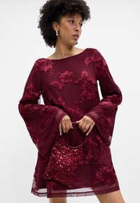 Βουργουνδί φόρεμα με λουλουδάτη κεντητική και κουβέντες μανίκια, συνδυασμένο με μια ταιριαστή τσάντα από πούλιες με κυκλικό χερούλι.