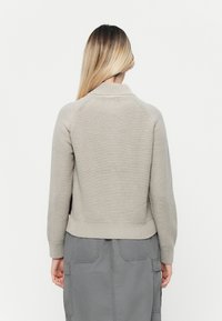 Beige gebreide sweater met een ribbelkraag en -manchetten, met een getailleerde taille en raglanmouwen. Draagt een grijze rok.