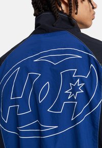 Modrá a černá bunda s velkým bílým logem na zádech. Vyrobená z lehké tkaniny s hladkou texturou a kontrastním prošitím.