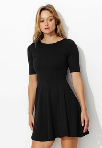 Robe noire à manches courtes, décolleté rond et jupe évasée. Fabriquée dans un tissu lisse avec des coutures subtiles et un corsage ajusté.
