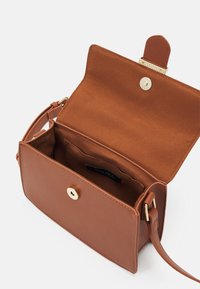 Sac bandoulière en cuir marron avec fermeture dorée, rabat ouvert montrant une poche intérieure et une étiquette de marque, bandoulière réglable attachée sur les côtés.