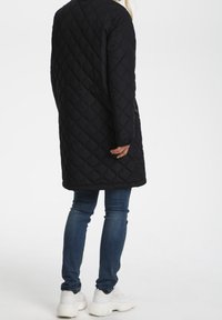 Abrigo largo negro acolchado con patrón de diamantes, de corte recto, detalles de botones, combinado con vaqueros azules y zapatillas blancas.