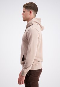 Bež hoodie iz mehkega materiala, z kenguru žepom, kapuco s vrvico in rebrastimi manšeti. Nosi se z rjavimi teksturiranimi hlačami.