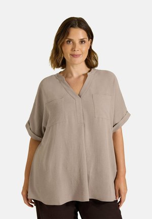 Mujer con cabello castaño hasta los hombros, llevando una blusa beige suelta de manga corta con bolsillos delanteros y pantalones oscuros contra un fondo liso.