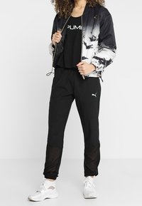 Svart bomberjacka med vit gradation och abstrakt mönster, kombinerad med en svart linne, svarta byxor och vita sportskor.