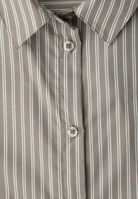 Koszula na guziki w jasnobrązowym kolorze z pionowymi białymi paskami. Posiada spiczasty kołnierzyk, zapięcie na guziki oraz gładką teksturę.