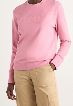 Personne portant un sweat-shirt rose à manches longues avec le texte "BOSS" et un pantalon beige, debout avec une main dans la poche devant un fond uni.