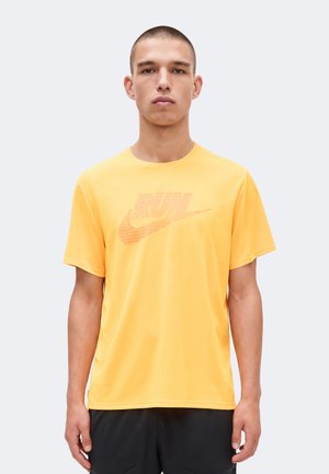 Jeune homme debout portant un T-shirt jaune Nike « RUN » et un short noir sur un fond clair uni.