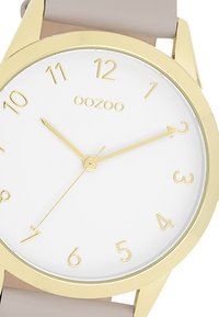 Gold-tone wristwatch with a white dial, Arabic numerals, and slender gold hands. Soft beige silicone strap. Minimalist design.