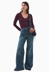 Donna che indossa un top viola a maniche lunghe con scollatura a V, jeans blu a vita alta e gamba larga, scarpe nere e una borsa a spalla nera.