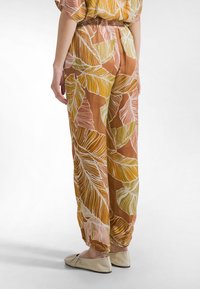 Brązowe spodnie z dużymi wzorami liści w kolorach żółtym, różowym i białym. Lekka tkanina z elastycznym pasem i marszczonymi mankietami. Noszone płaskie buty.