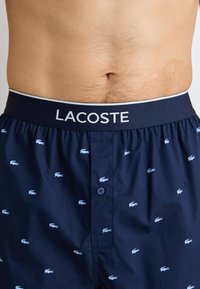 Navy blue boxer shorts with a logo waistband, featuring a repeated small light blue crocodile pattern and a front button closure.