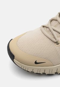 Beige atletiske sko med tekstureret stof, rund tå og en sort gummisål. Har snørebånd og et lille sort Nike-logo.