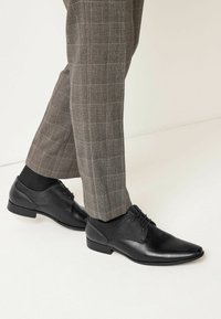 Zapatos de vestir de cuero negro con un diseño elegante de punta afilada, que presentan texturas sutiles y cordones, combinados con pantalones a cuadros grises.