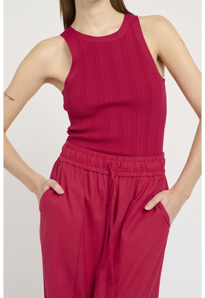 Femme portant un haut rouge sans manches côtelé et un pantalon rouge taille haute avec cordon de serrage, les mains dans les poches, sur un fond uni.