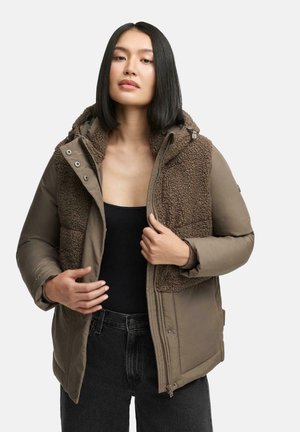 Bruine pufferjack met een fuzzy, gestructureerd bruine fleece voorkant, ritsluiting en capuchon. Draagt over een zwart topje en jeans.