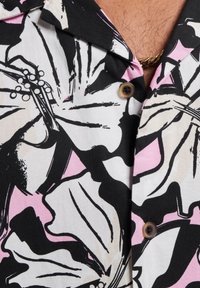 Camisa de manga corta con un patrón floral en negro, blanco y rosa. Cuenta con un cuello y un botón marrón, hecha de tela ligera.