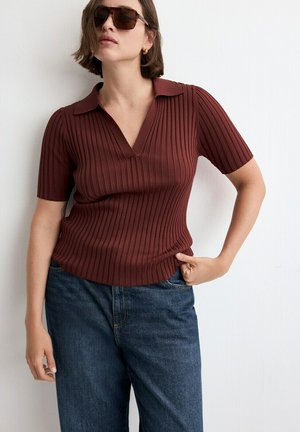 Žena v tmavě červeném žebrovaném polotriku s krátkými rukávy, tmavých slunečních brýlích, zlatých kruhových náušnicích a modrých džínách stojí u bílé stěny.