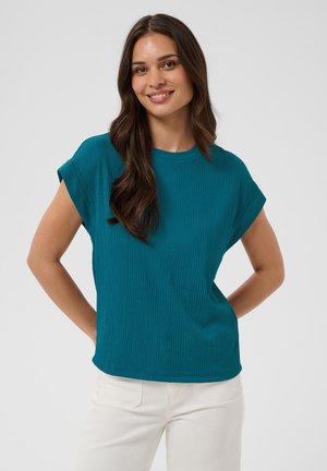 Jeune femme aux longs cheveux bruns portant un haut bleu canard à manches courtes et un pantalon blanc, souriante, les mains derrière le dos.