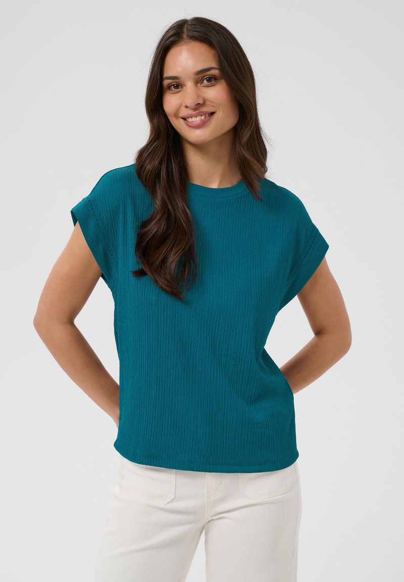 Jeune femme aux longs cheveux bruns portant un haut bleu canard à manches courtes et un pantalon blanc, souriante, les mains derrière le dos.