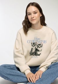 Felpa beige con una grafica di due gattini, testo "COORDINATORI DEL CAOS" e polsini a coste. Indossata con jeans blu.