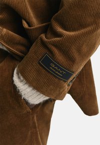 Manche de manteau en velours côtelé brun avec une finition texturée et une étiquette GANT bleu foncé. Accentuée par un tissu léger qui dépasse en dessous.
