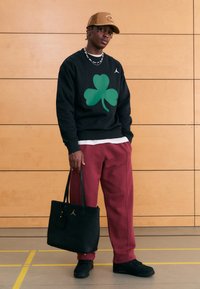 Svart genser med stort grønt kløveremblem, røde joggebukser, svart totebag med gulllogo og brun caps. Bakgrunn av trepanel.