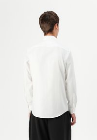 Camicia bianca a maniche lunghe con colletto classico, texture liscia, polsini con bottoni e orlo arrotondato, indossata con pantaloni neri.