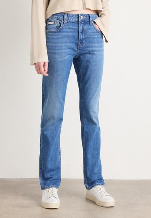 Osoba ubrana w jasnoniebieskie proste dżinsy, beżową krótką koszulę z długim rękawem, białe trampki, stojąca na jednolitym tle.