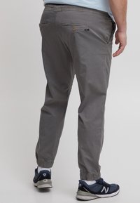 Pantaloni sport gri dintr-un material ușor. Manșete elastice la glezne, două buzunare posteriori și detalii cu etichetă de marcă lângă talie.
