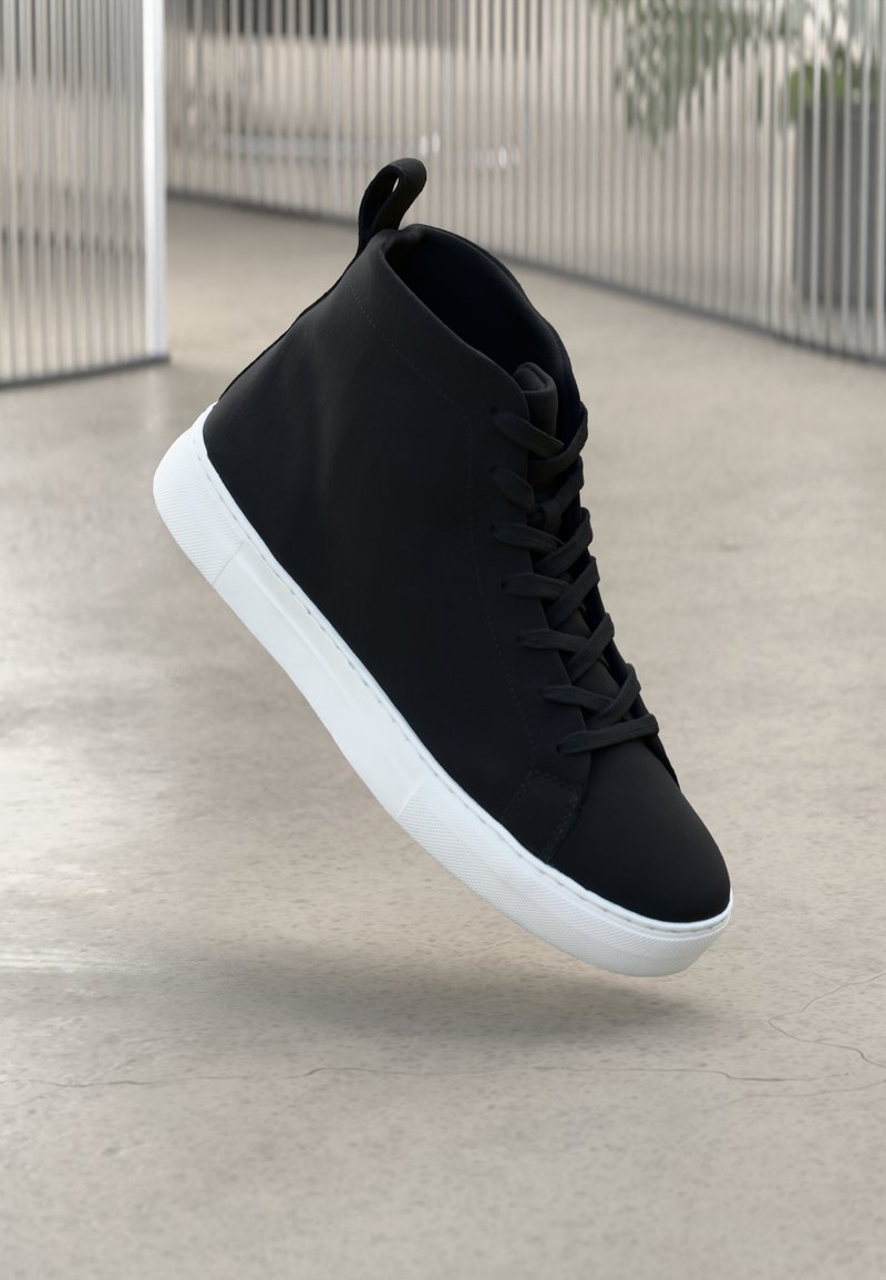 Sorte høyhælte sneakers med en glatt lærfinish, flat hvit gummisåle og svarte snørebånd. Rund tå og hylse bak.