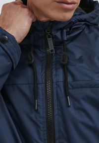 Tamsiai mėlynas užtrauktuku užsegamas džemperis su kapišonu, su juodais raišteliais ir detalėmis, lygaus paviršiaus, ir priekinėmis kišenėmis.
