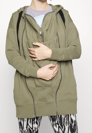 Osoba ubrana w oliwkową bluzę z kapturem na suwak z dwoma otwartymi suwakami, trzymająca tkaninę wystającą z przedniej kieszeni bluzy.