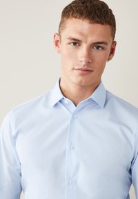 Svetlomodrá košeľa s jemnej látky, s klasickým golierom, zapínaním na gombíky a jemnou textúrou. Štandardný strih s upraveným vzhľadom.