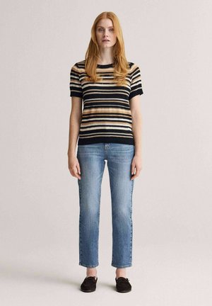 Nainen, jolla on pitkät punaiset hiukset, pukeutuneena mustaan, ruskeaan ja valkoiseen raidalliseen lyhythihaiseen neulepaitaan, vaaleansiniset farkut ja mustat mokkasiinit.