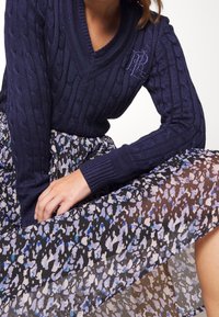 Navy cable-knit sweater with V-neck and logo, paired with a semi-transparent skirt featuring a blue and black abstract pattern.