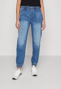 Světle modré joggingové kalhoty z denimu, s páskem na šňůrku, elastickými manžetami a volným střihem. V kombinaci s bílými sportovními botami.
